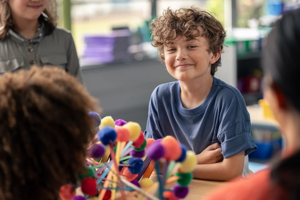 A boy with curly hair smiles in a classroom setting, seated at a table with colorful, handmade objects, surrounded by other children.