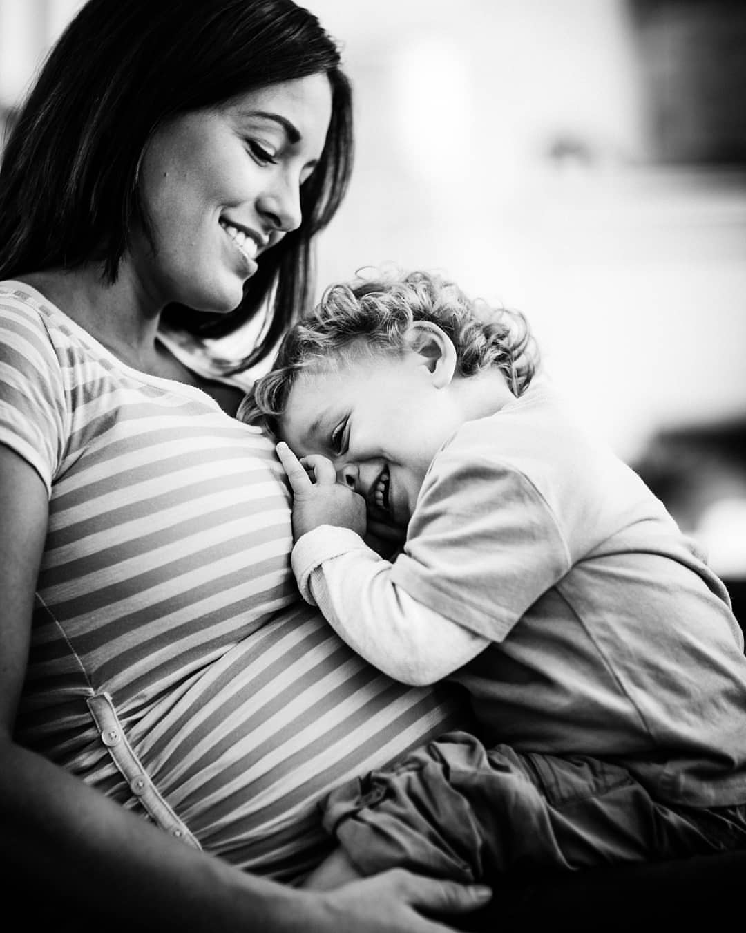 Black and white photo of a pregnant woman in a striped shirt with a happy child leaning on her stomach in an indoor setting.
