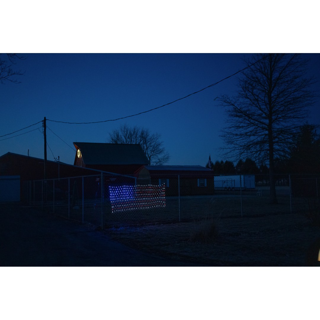 Illuminated American flag display hangs on a fence outside a rural home at dusk with a red barn and trees in the background.