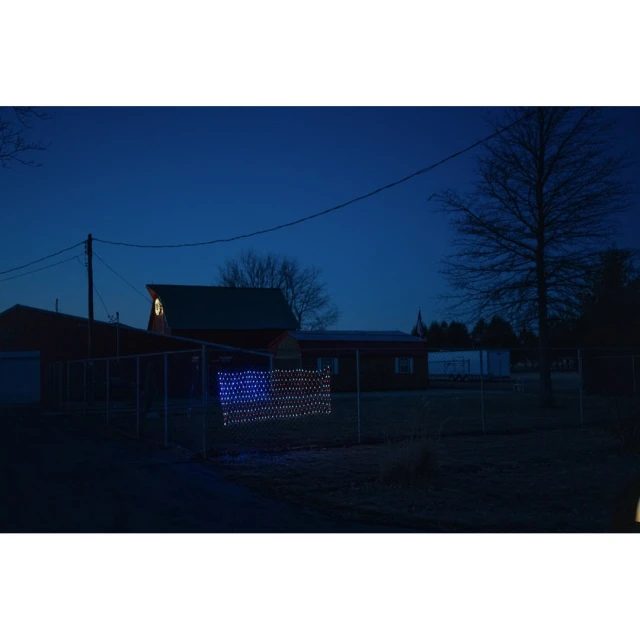 Illuminated American flag display hangs on a fence outside a rural home at dusk with a red barn and trees in the background.