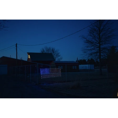 Illuminated American flag display hangs on a fence outside a rural home at dusk with a red barn and trees in the background.
