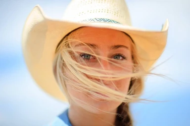 Close-up portrait of a woman wearing a straw cowboy hat with blonde hair blowing across her face outdoors.
