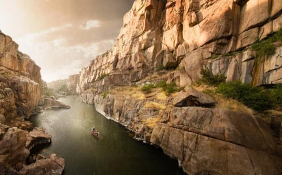 A small canoe with people navigating a dark river carved through steep rocky canyon walls under a glowing overcast sky