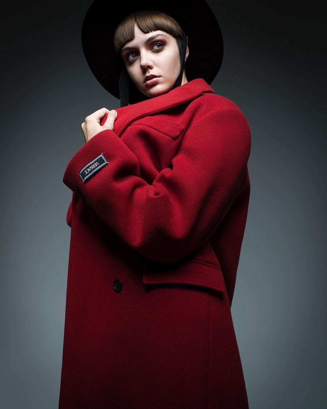 Stylish female model in a red coat and black hat posing in a studio setting. Fashion editorial shot with a modern aesthetic.