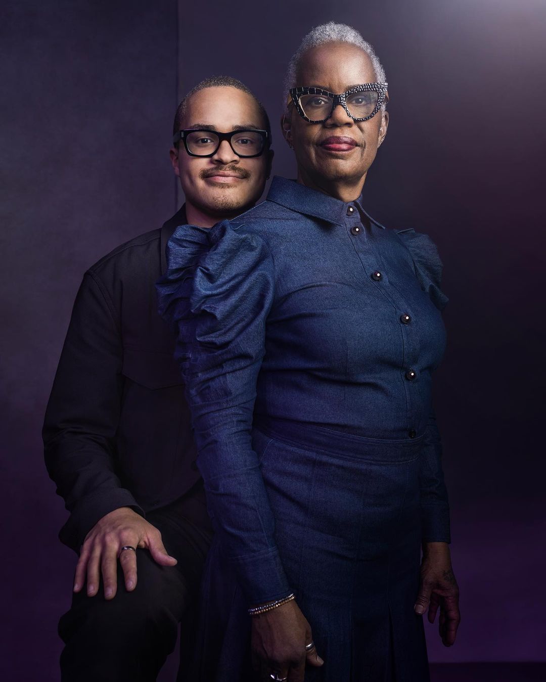 Portrait of a mother and son in a studio setting. The woman is wearing glasses and a blue jean dress. The man is wearing a black shirt and glasses.