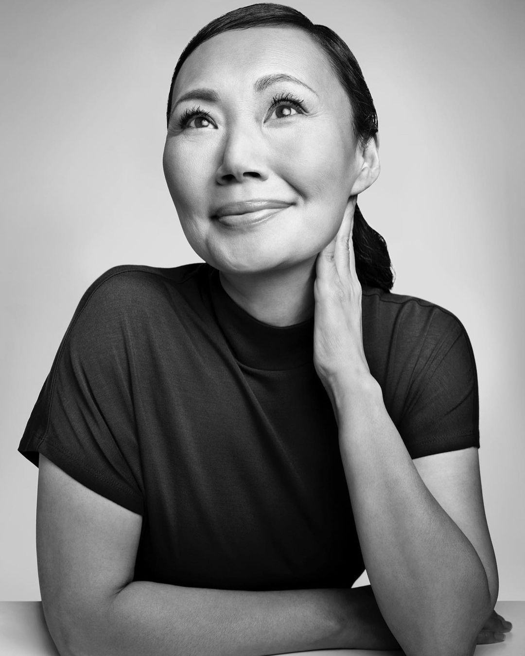 Black and white portrait of an adult woman in dark top against a neutral background, hand touching neck, looking up and to the side.