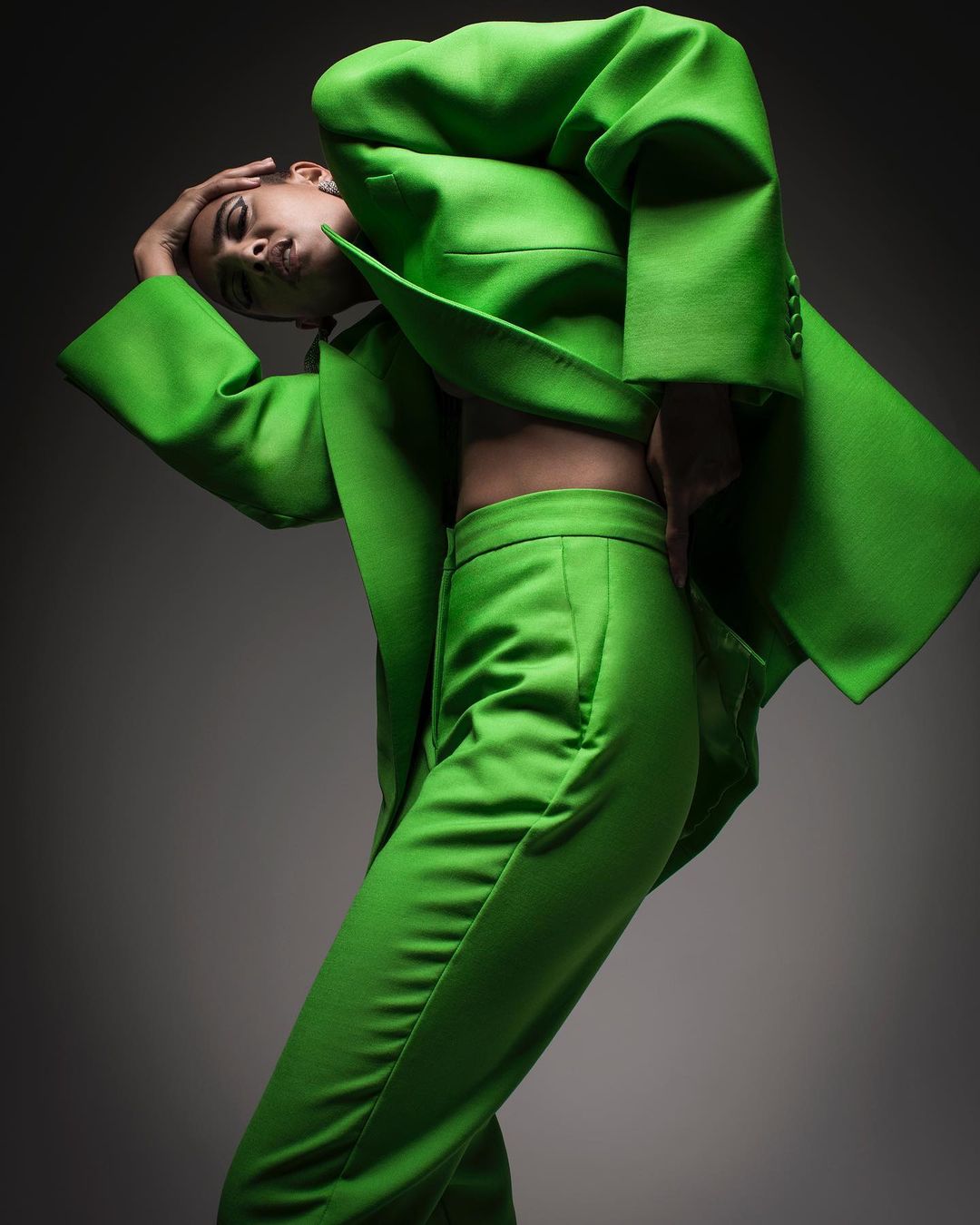 A fashionable female model poses in a vibrant green suit against a studio backdrop, showcasing a modern and stylish aesthetic.