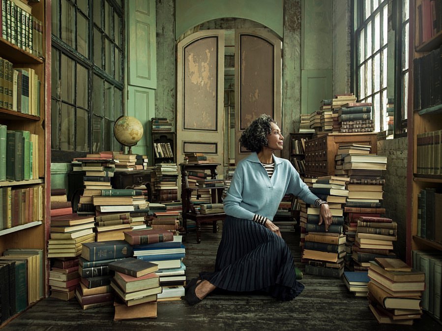 Adult woman sits among a collection of books in an interior scene with large windows and doors, wearing casual clothing in soft light.