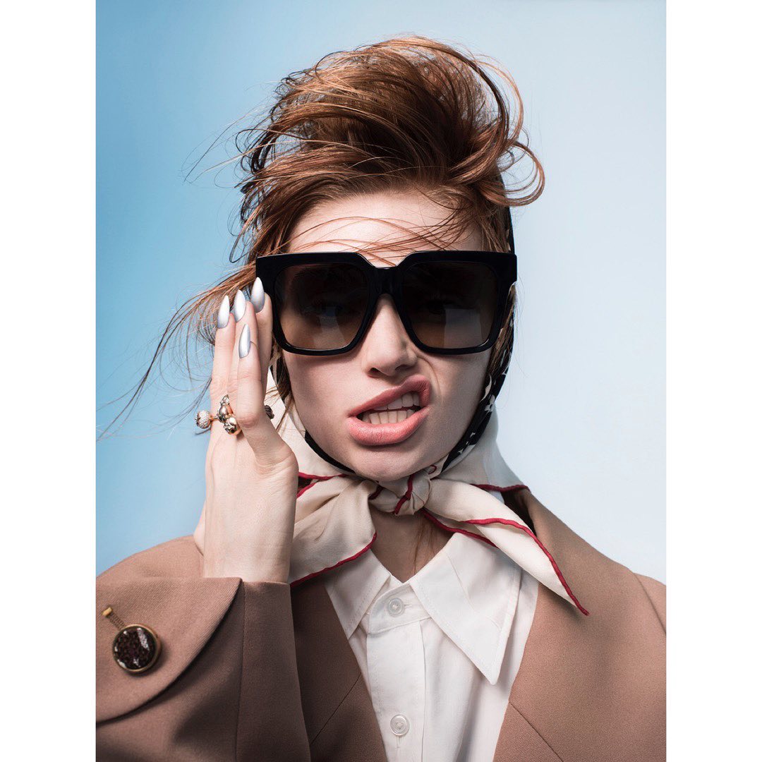 Portrait of a redhead woman in sunglasses, wearing a tan blazer and headscarf against a gradient blue background in a studio setting.