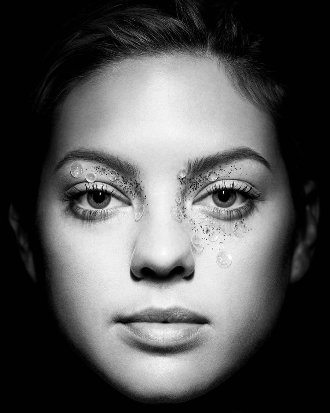 Close-up black and white portrait of a woman with glitter and droplets around her eyes, set against a dark background.