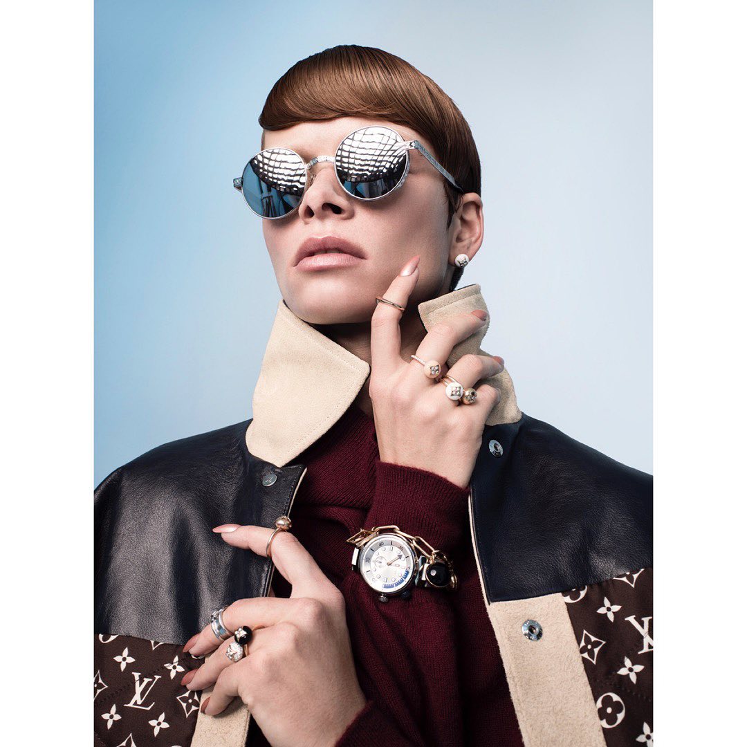 Fashion shot of a woman wearing reflective round sunglasses, rings, and a watch, against a light blue background in a studio.