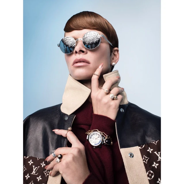 Fashion shot of a woman wearing reflective round sunglasses, rings, and a watch, against a light blue background in a studio.