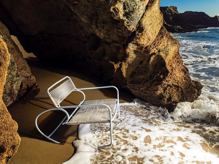 A modern white armchair sits on a sandy beach, partially submerged by ocean waves, framed by large, textured rocks under a clear blue sky.