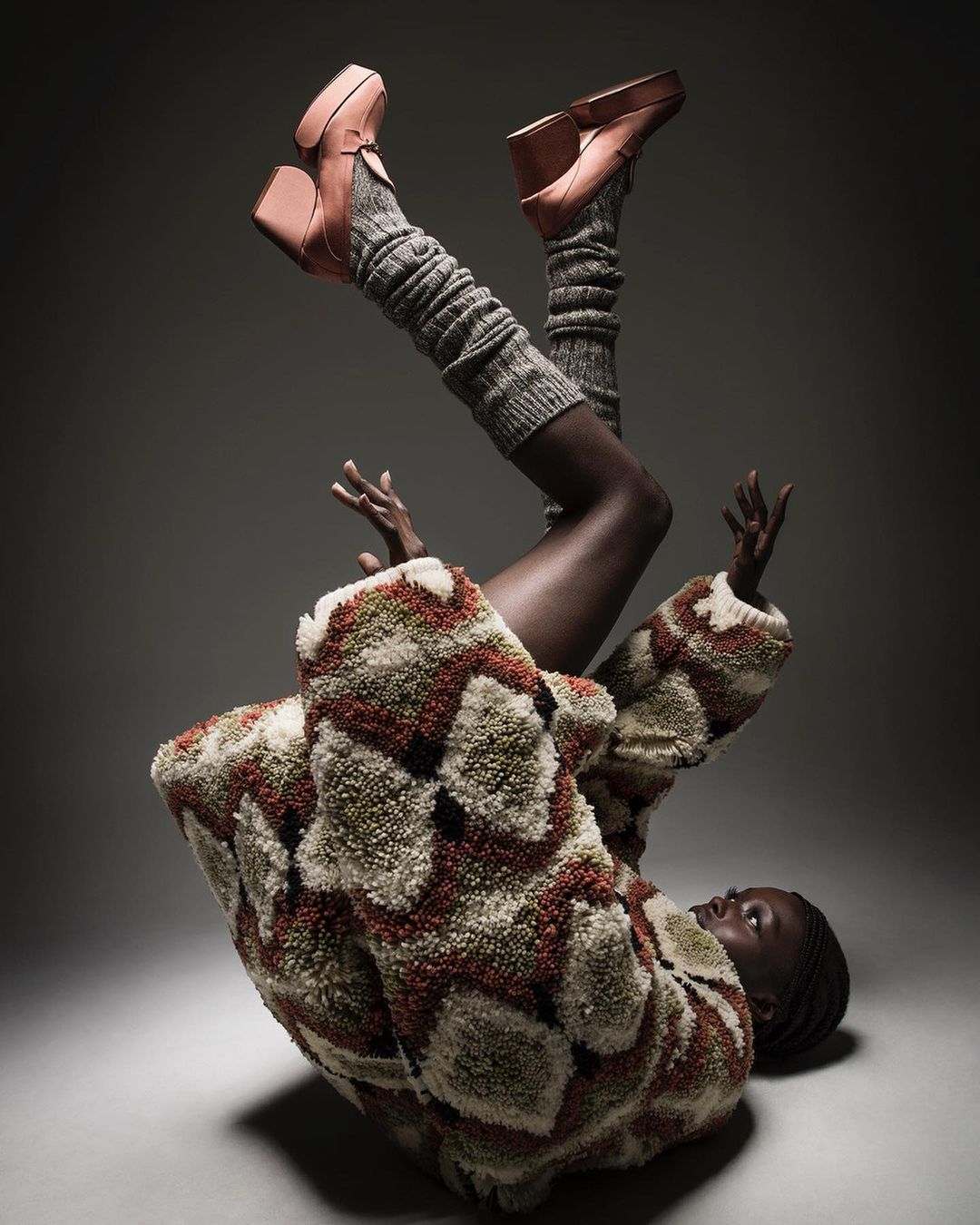 A Black woman in an upside-down pose with platform heels, leg warmers, and a patterned sherpa jacket against a gray background.