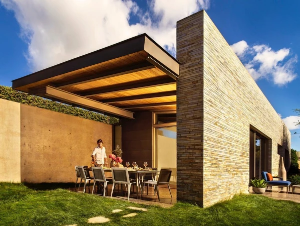 Modern house with stone exterior, patio table set for dining, and person setting up. Grass lawn in foreground, blue sky above.