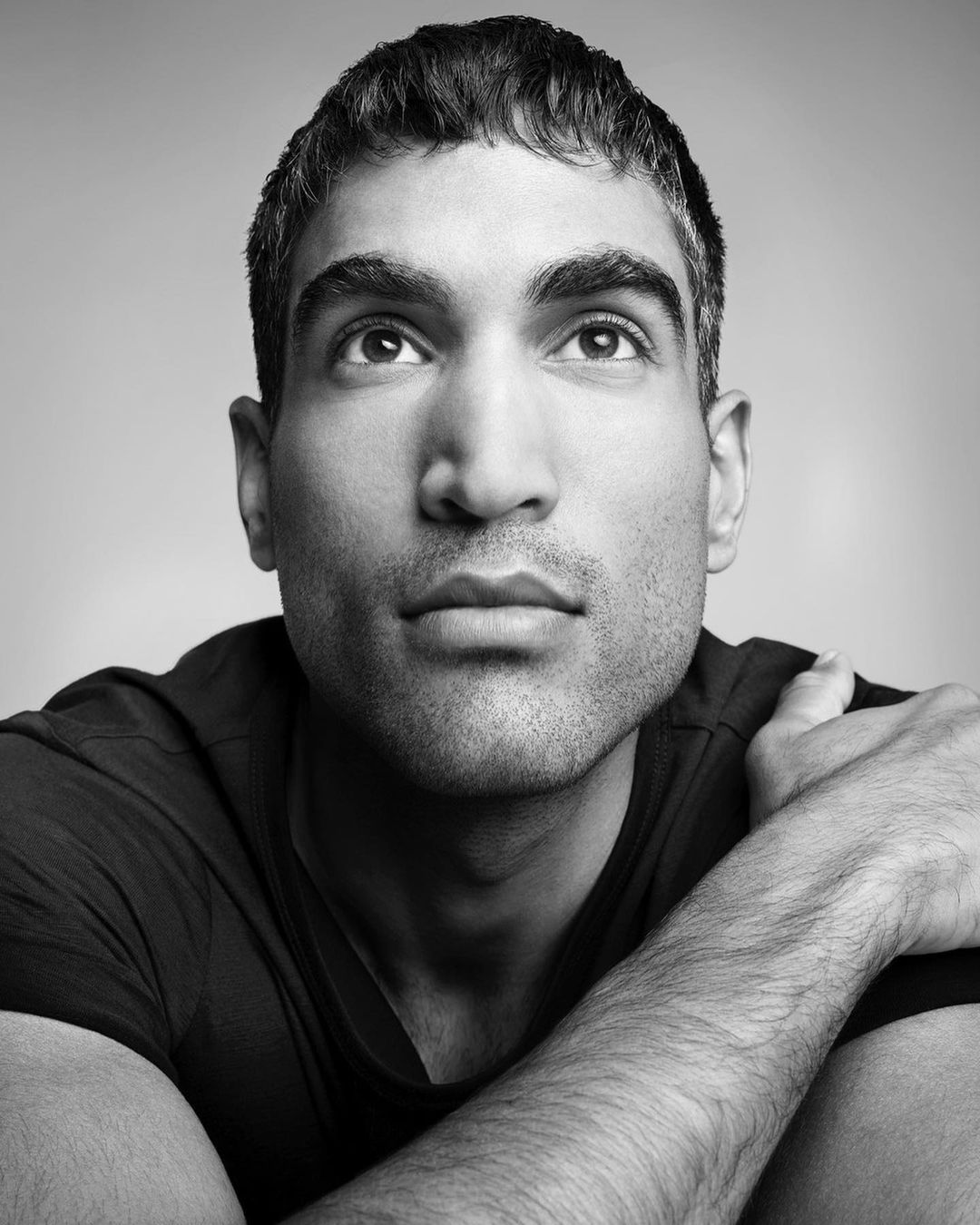 Black and white portrait of a man with dark hair and eyes looking up, arms crossed over his chest against a light gray background.
