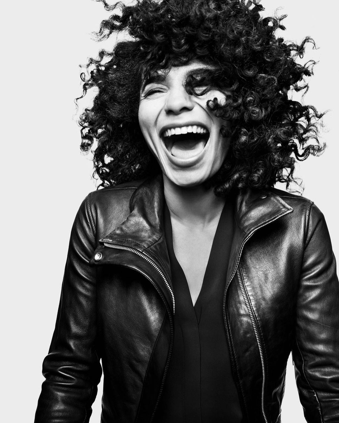 Black and white portrait of a smiling woman with curly hair wearing a leather jacket against a neutral background, emphasizing her joy.
