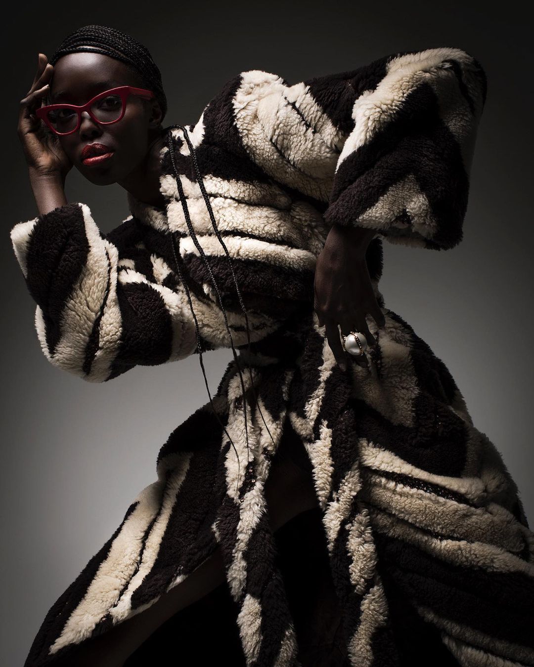 Fashionable dark-skinned woman in bold zebra-striped coat and red glasses, striking a pose against a neutral gradient backdrop.