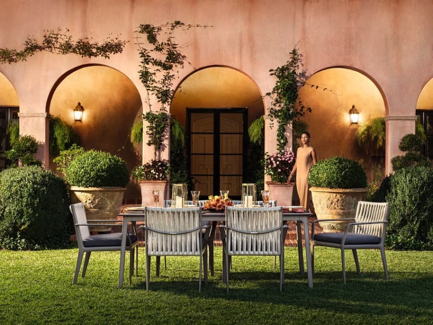 Outdoor dining area with a table, six chairs, and a woman standing in the background in front of an archway in a garden setting.