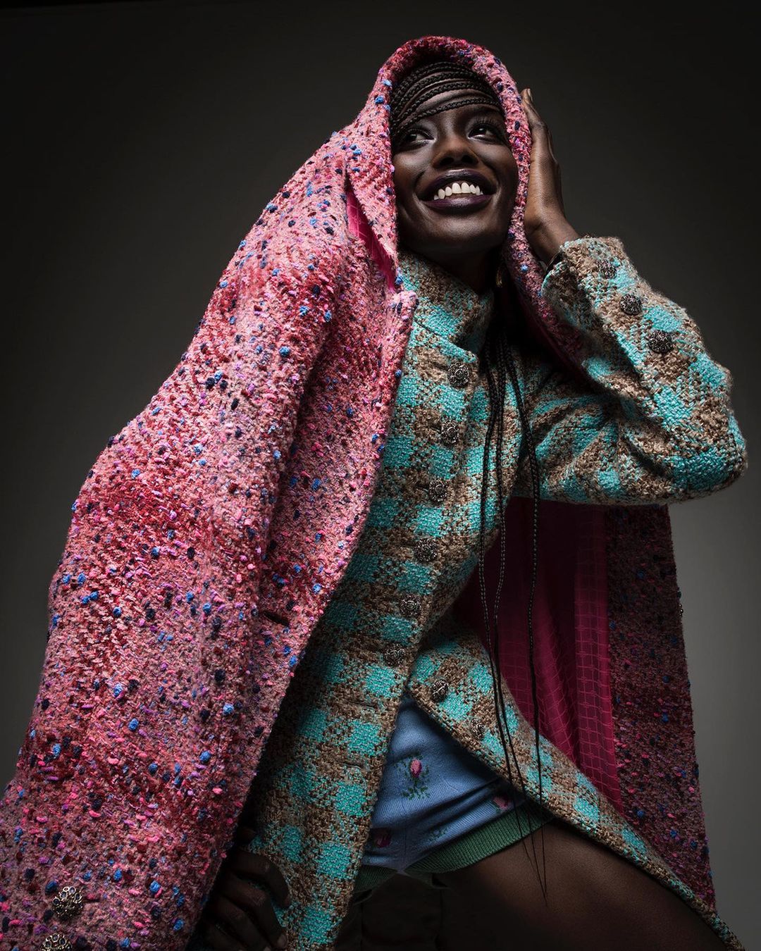 Smiling African woman in an ornate, patterned coat and hood, photographed in a studio with a neutral grey backdrop and soft lighting.