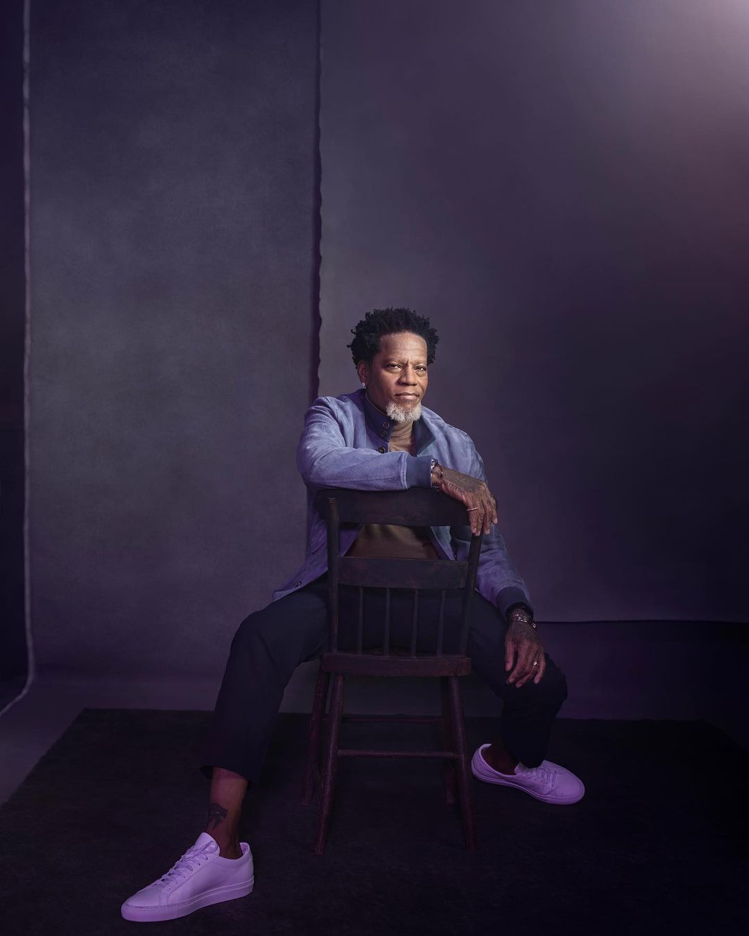 Portrait of an adult male sitting backwards on a wooden chair in a photo studio with a gray backdrop, wearing a blue shirt and white sneakers.