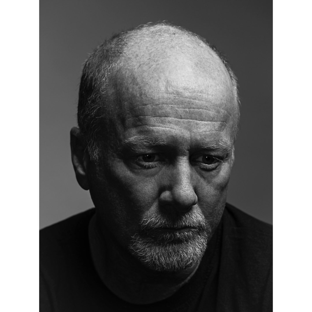 Black and white portrait of a serious adult male with a bald head, light beard, and dark t-shirt against a plain grey background.
