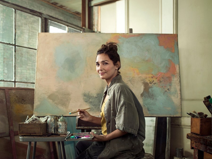 Woman artist sits indoors in front of a large abstract painting, holding a brush and a paint palette, in a brightly lit art studio.