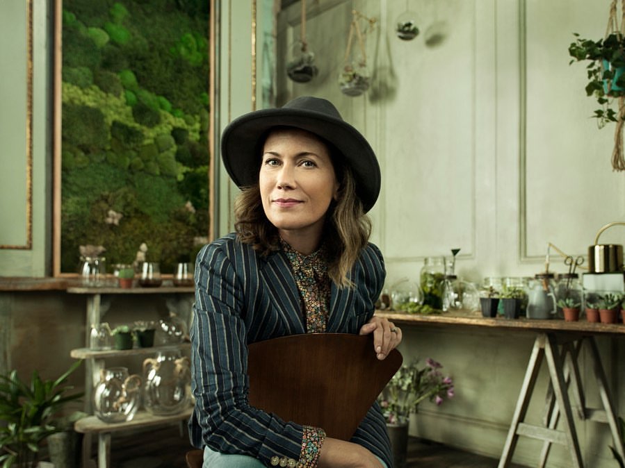 Adult woman in a hat and pinstripe blazer poses indoors in front of plants and glass jars on a shelf, in a portrait shot.