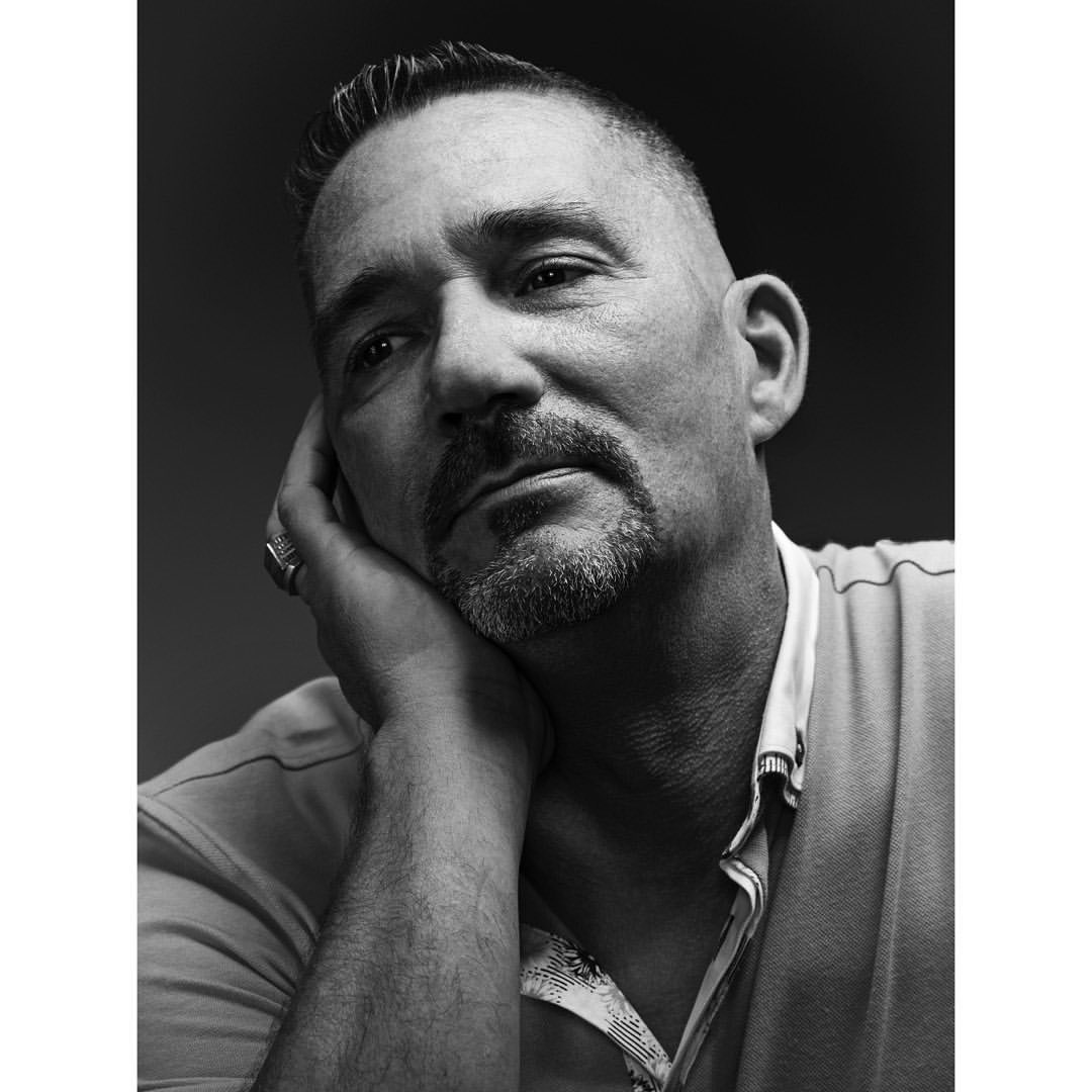 Black and white portrait of a middle-aged man with hand on face. Man has short hair, a goatee and is wearing a collared shirt.