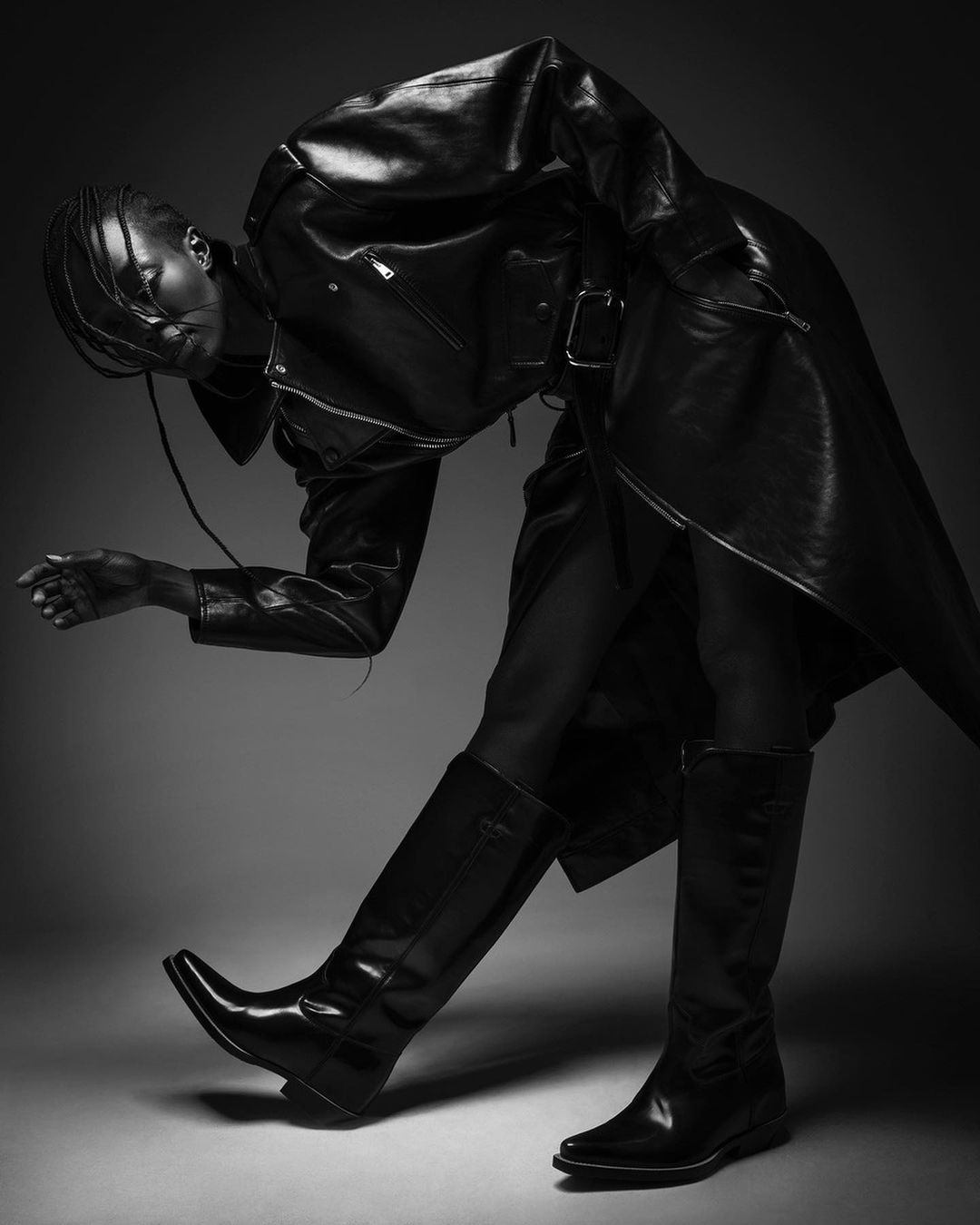 Black and white studio shot of a Black female model wearing a black leather jacket and boots, with a neutral gray background.