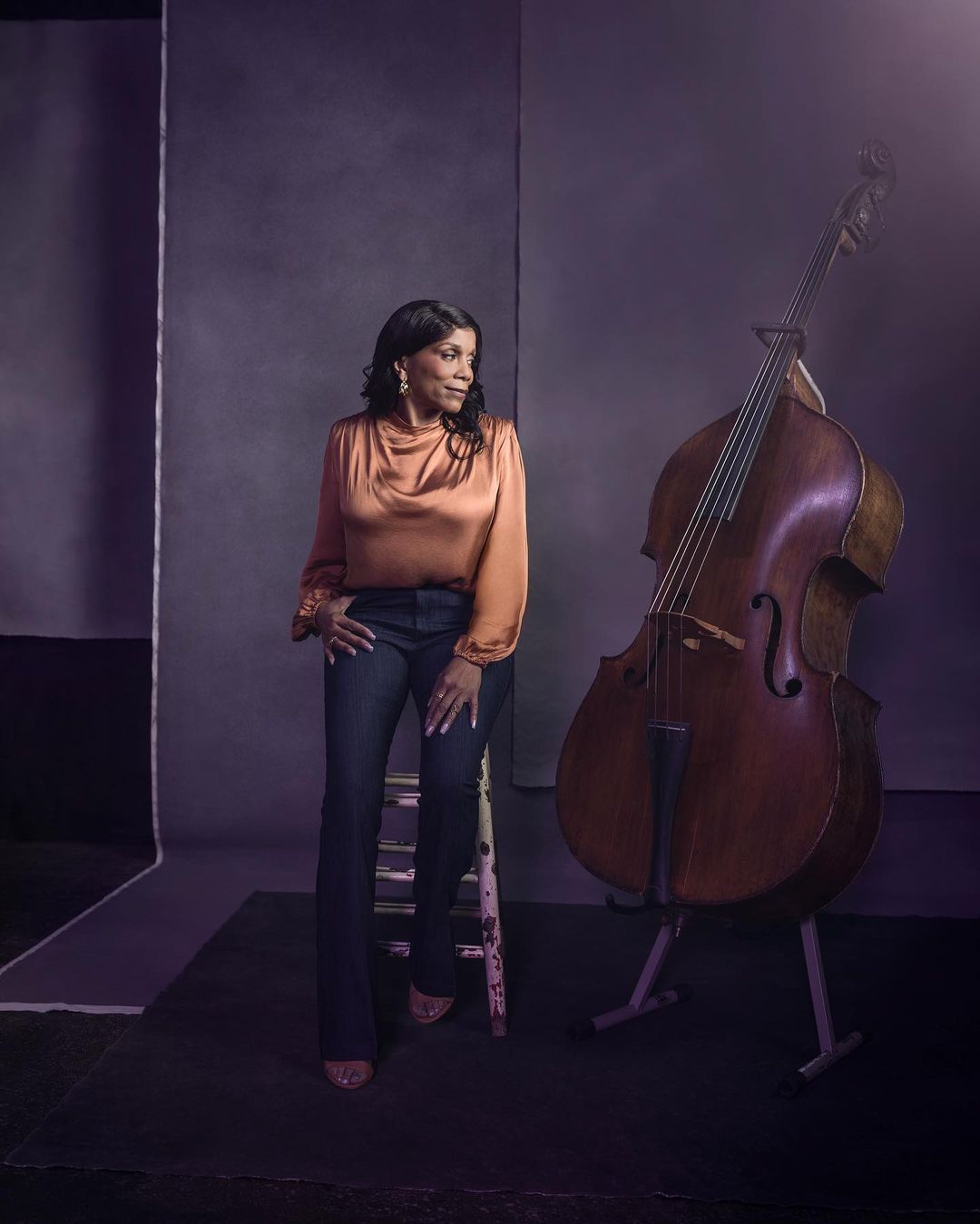 Portrait of an adult woman seated on a stool next to a cello in a photo studio, gazing away from the camera with a serene expression.