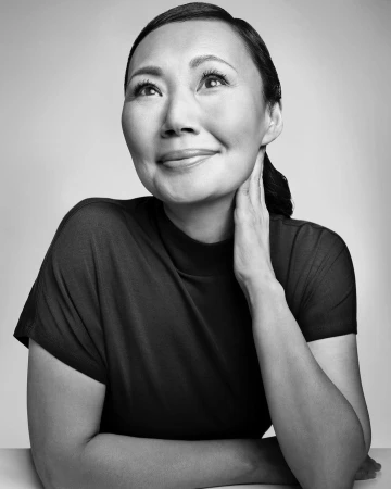 Portrait of an East Asian woman with dark hair wearing a dark mock neck top, smiling upward against a light background.