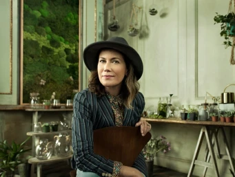 A woman in a grey fedora and striped blazer seated indoors against a green wall filled with moss and plants.