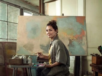 Female artist holding paintbrush and palette sitting before a large canvas in a bright art studio with large windows.