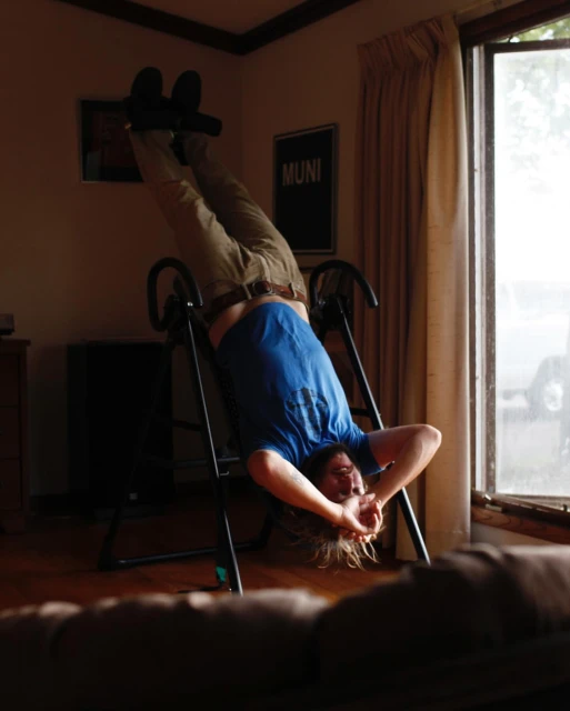Adult male using an inversion table inside a room with window, tan curtains, and a Muni poster on the wall; indoor fitness equipment.