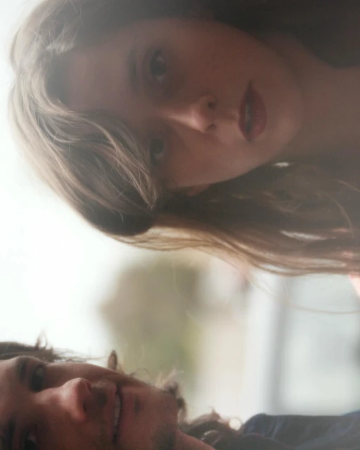 Eye-level shot of a young adult woman and man, faces close together, expressing connection in a softly-lit, hazy, outdoor setting.