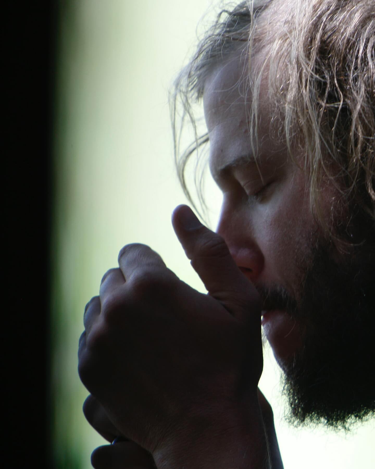Portrait of adult male with a beard, indoors, eyes closed, and hands clasped against his chin in a contemplative or meditative pose.