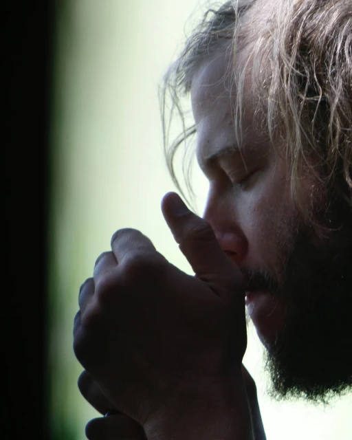 Portrait of adult male with a beard, indoors, eyes closed, and hands clasped against his chin in a contemplative or meditative pose.