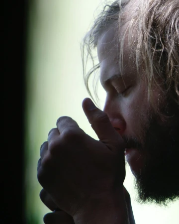 Portrait of adult male with a beard, indoors, eyes closed, and hands clasped against his chin in a contemplative or meditative pose.