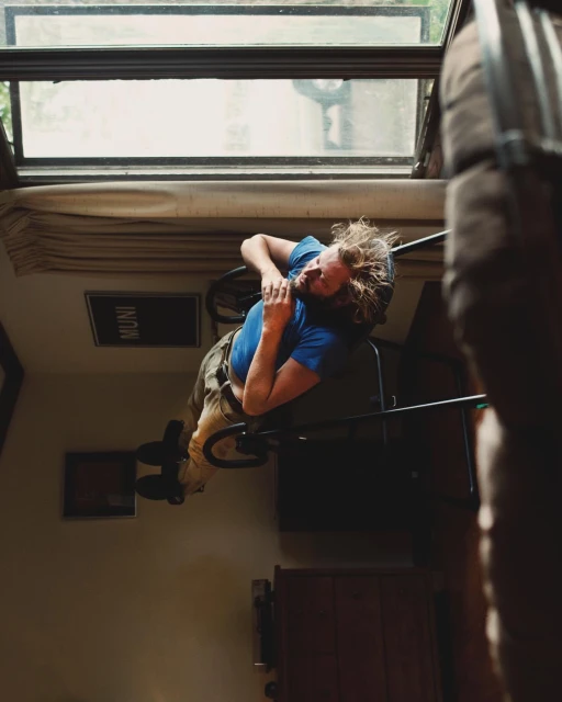 Man lying back in a chair indoors with an overhead window and muted color palette, creating a tranquil, artistic composition.