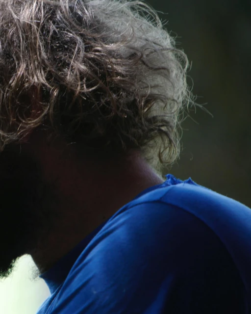 Close-up of an adult with graying hair wearing a blue shirt, facing away from the camera in an outdoor setting, focusing on hair and shoulders.