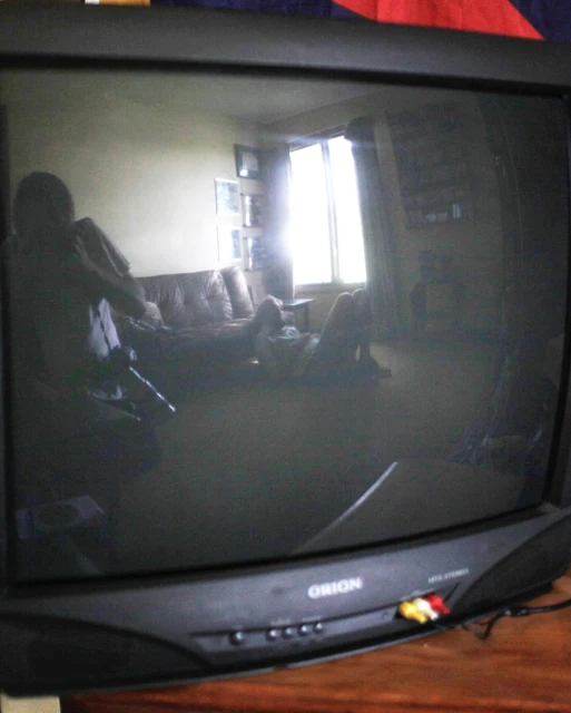 Old CRT television reflecting interior scene with couch, window, and figures, set against a dark wood surface.