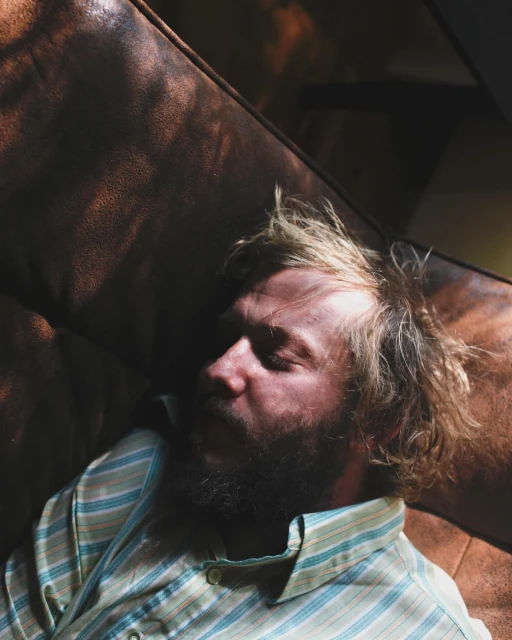 Bearded man in a striped shirt lies asleep on a brown leather couch in the light, close-up shot, providing a candid and intimate glimpse.