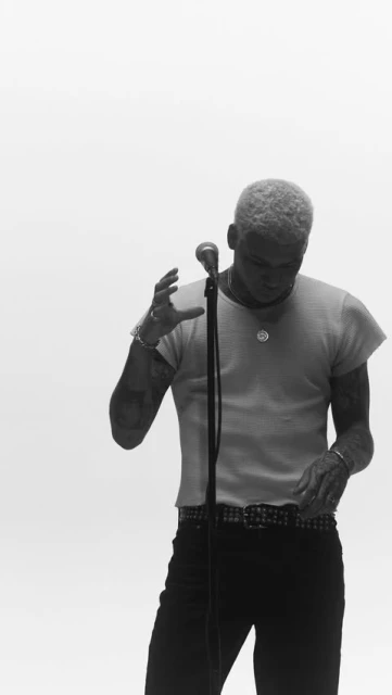 A man with fair hair stands in front of a microphone, wearing a white t-shirt, dark pants, and a studded belt in a studio environment.