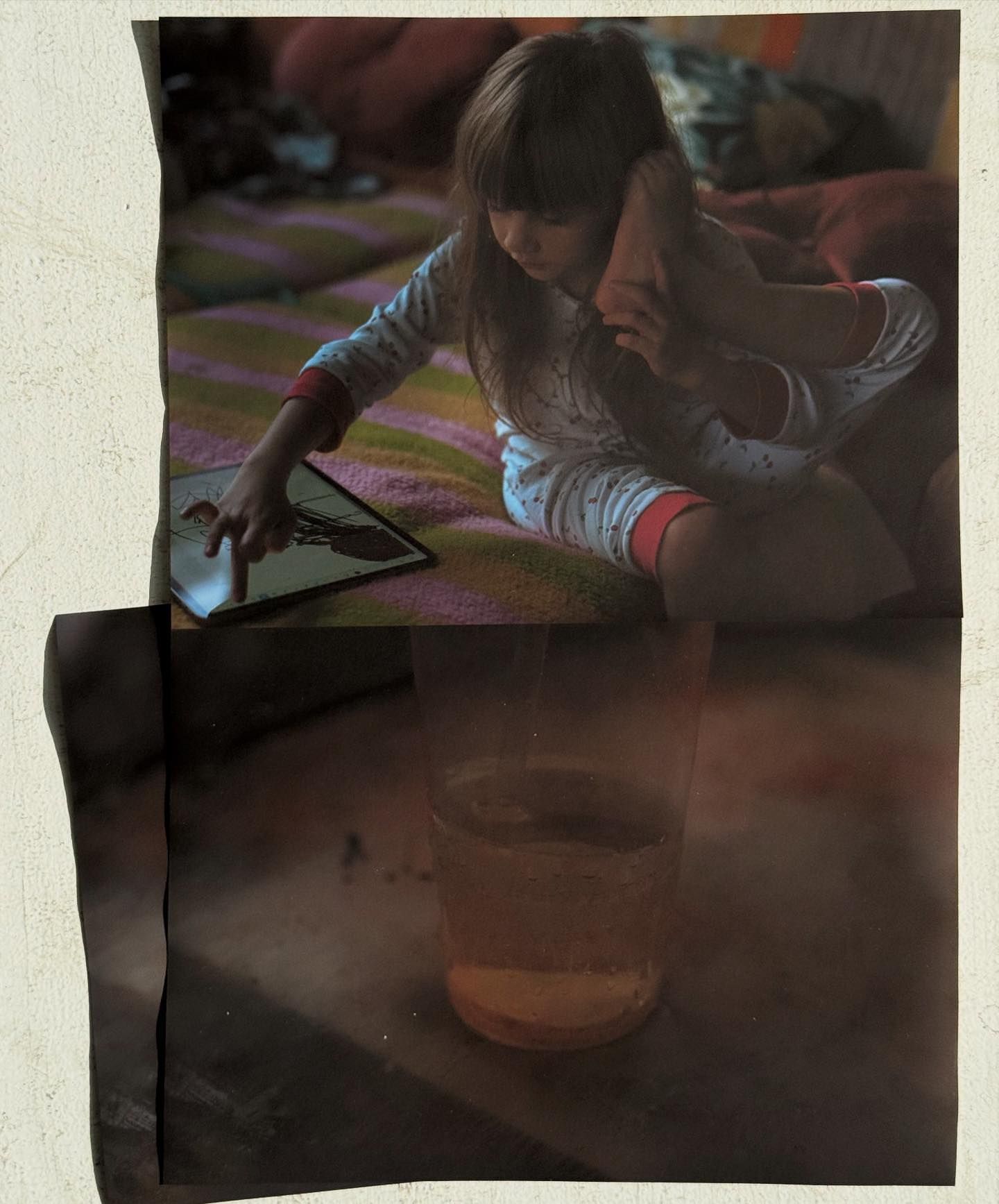 Young child with long hair wearing pajamas sits inside using a tablet; a glass of liquid is visible in the foreground.