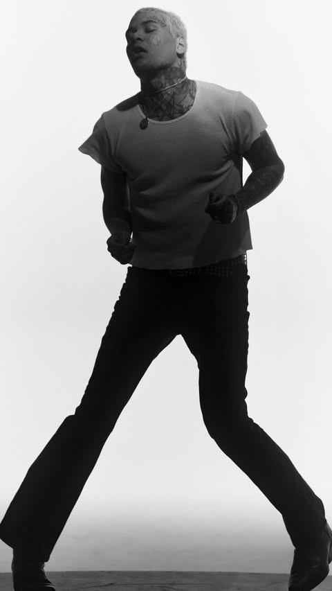 Monochromatic portrait of a person with short blonde hair, tattoos, t-shirt, and dark pants against a white backdrop in studio lighting.
