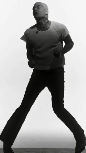 Monochromatic portrait of a person with short blonde hair, tattoos, t-shirt, and dark pants against a white backdrop in studio lighting.