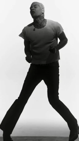 Monochromatic portrait of a person with short blonde hair, tattoos, t-shirt, and dark pants against a white backdrop in studio lighting.