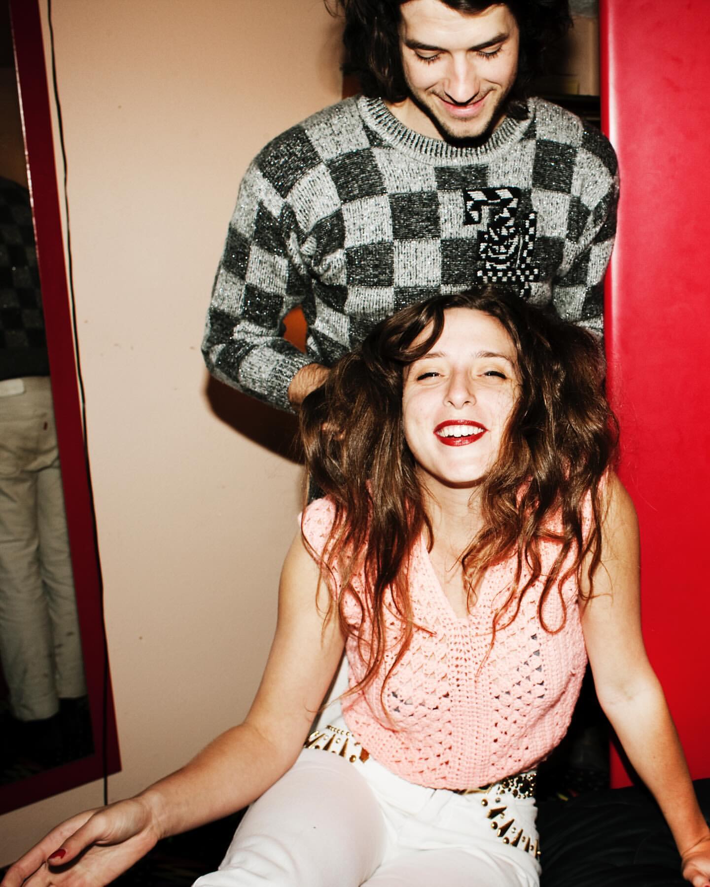 A happy couple posing indoors. A woman with curly hair sits while a man with long hair stands behind her. They smile in a casual setting.
