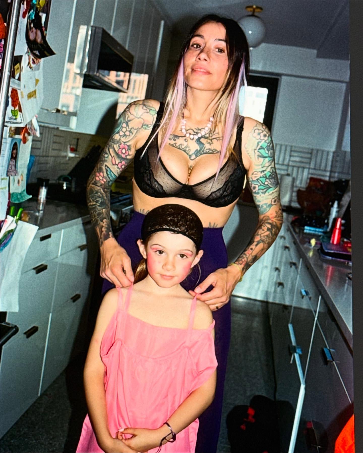 Woman and child pose indoors. Woman has tattoos and a black bra. The child wears pink dress. The scene is domestic and personal.